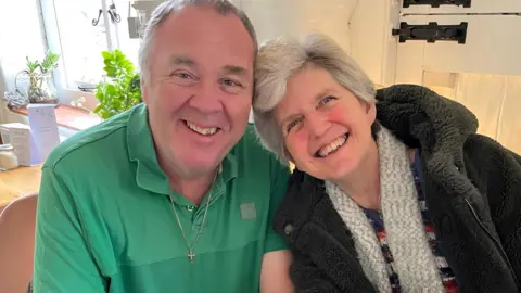 A couple sitting and smiling at the camera. The man is wearing a green polo shirt and a gold cross necklace, while the woman is dressed in a dark jacket, colourful top, and a light grey knitted scarf. There's a window and a plant behind them.