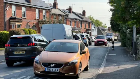 Busy street scene on Church Street, Leckhampton, Cheltenham, Gloucestershire. Several cars on road, street lined with terraced houses. 
