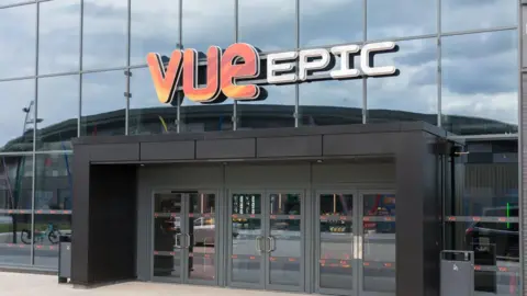 The Vue cinema building displays the Vue EPIC branding. The building has a large six-door entrance and large glass windows.