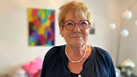 A woman wearing glasses stands in her living room and smiles at the camera.