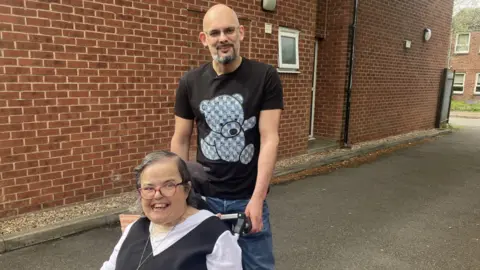Luke is taking one of the residents Emma out for some fresh air in her wheelchair. She is smiling and he is pushing the wheelchair outside. 