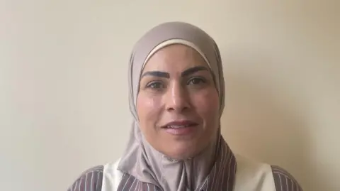 Wearing head scarf, striped shirt and white waist coat. Standing in front of a white wall.