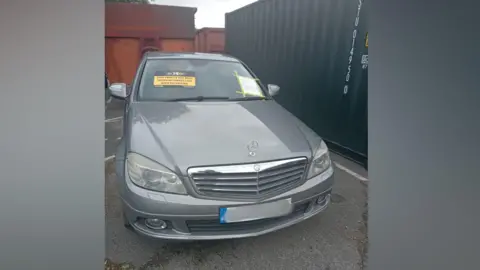 A grey Mercedes Benz car with its registration plate blurred is parked next to a shipping container. It has a yellow sticker on it which reads 'This vehicle has been seized in connection with fly-tipping'.