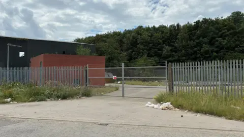 A small brick building is behind a barbed wire gate and metal fence. There is rubbish blown against long grass near the entrance.