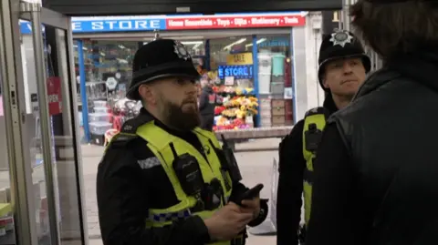 PC Gareth Bielby on the left and Sergeant Lee Waller on the right are standing in front of a man as they question him. Both officers are wearing police uniform including a yellow high vis vest with a body cam and radio attached. They are both wearing police hats. They are all inside a shop and standing close together. The man in front of the officers is dressed in black and has brown hair.