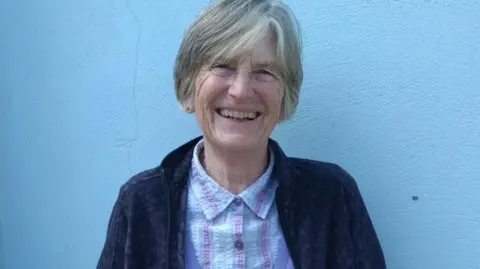 MANX DEAF SOCIETY Lesley Cullen, a woman with short grey hair, she has a friendly smile and wears a blue shirt and navy jacket, and is stood in front of a blue wall.