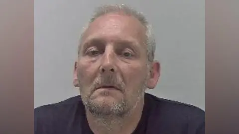 NCA A mugshot of an older man with grey hair and grey facial hair. He is wearing a dark navy t-shirt