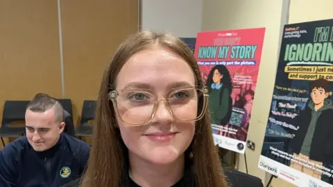 Neve Robinson, a woman with long brown hair, who is wearing large framed glasses.