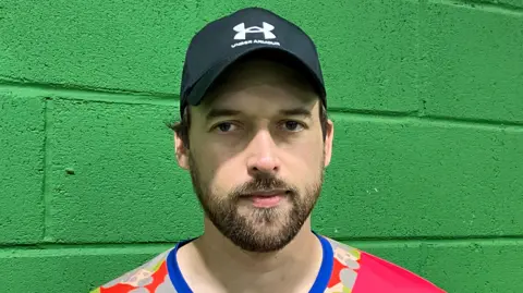 A man in a baseball cap and pink t-shirt with a beard standing in front of a green brick wall 