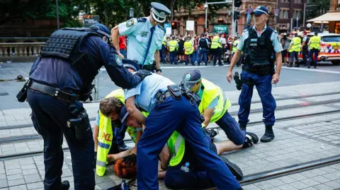 Getty Police officers in blue uniforms hold down a person on a city street with a crowd of people and a police car in the background.