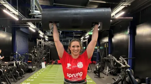 Luke Deal/BBC A woman in a gym raises a long cylinder-shaped weight above her head. She is smiling. She is wearing a red T-shirt. Various pieces of gym equipment are behind her. 
