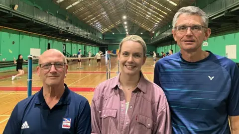 Dermott Heslop, Heather Robertson and Mike Poulter stand side by side in front of the Badminton terrains, where players can be seen a match. Mr. Heslop wears glasses and a naval sports top with a British judo logo on the chest, Mrs. Robertson wears a pink shirt and Mr. Poulter has a navy blue sport and glasses.