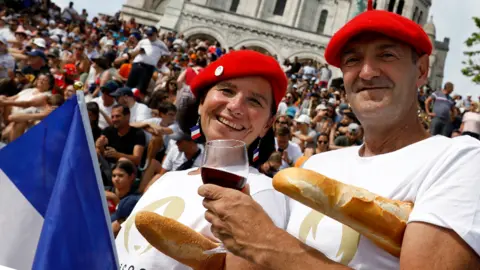 Getty Images Mężczyzna i kobieta w francuskich beretach trzymają przed tłumem czerwone wino i bagietki