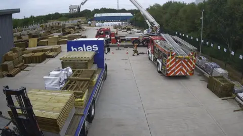 A concrete yard outside an industrial building. Piles of wooden pallets are on the trailer of a truck and on the ground on the left. Two fire engines are parked in the yard - one has its aerial appliance extended.