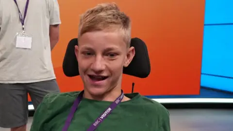 A young boy with short blonde hair sits in a wheelchair wearing a green t shirt.