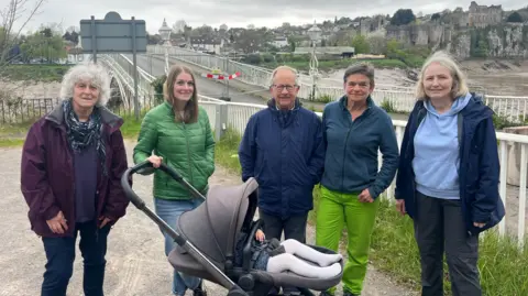 Image shows a group of people stand in a car park in front of a bridge, which is blocked by two barriers. The group consists of three women and two men, one of whom is with a pushchair. In the background, a river flows beneath the bridge, and beyond it stands an old castle.
