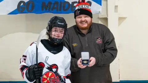 Alan Blakeley Caledonia Steel Queens head coach Richard 'Rambo' Gray presenting Molly, who is wearing a black and white hockey uniform and helmet with a medal for player of the match. Rambo is wearing a grey long-sleeved quarter zip top and a black and red bobble hat.
