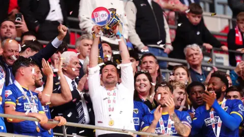 AFC Wimbledon manager Johnnie Jackson (centre), wearing a white shirt, lifts the trophy in the Wembley stands, with his players dressed in blue cheering and clapping beside him. There is confetti floating in the air.