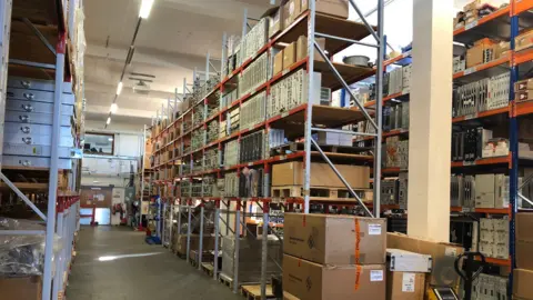 Martin Heath/BBC Large room showing three bays of metal racking, in which various spare parts of being stored. There are fluorescent lights suspended from the ceiling