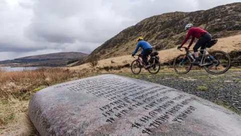 Two people on bikes cycle past a large stone with rugged hills in the background and a loch up ahead