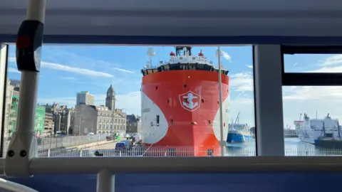 Rebecca Kerr A photo taken from inside a bus, looking out to a harbour (Aberdeen), with a large red and white oil industry vessel in view, as well as a NorthLink passenger ferry.