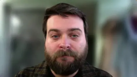 Dr Connor Bamford looking at the camera. He has dark brown hair, a moustache and a beard. He is wearing a green and dark brown checked shirt. The background behind him is blurred.