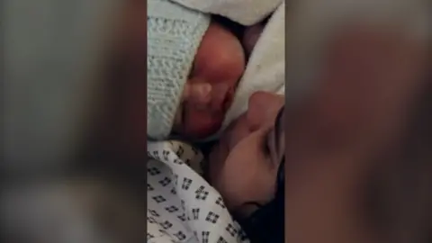 A mother with dark hair in a hospital gown, looking into the face of her baby just born by C-section.