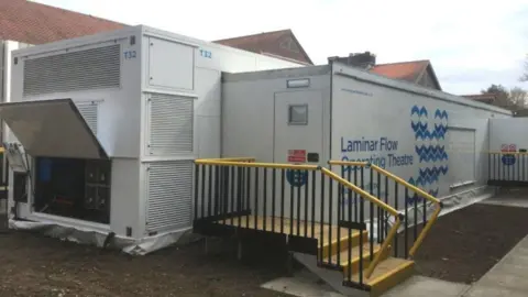 An example of the temporary wards. It is an outdoor mobile unit comprised of two metal containers joined together, with yellow stairs leading up to an entrance. 