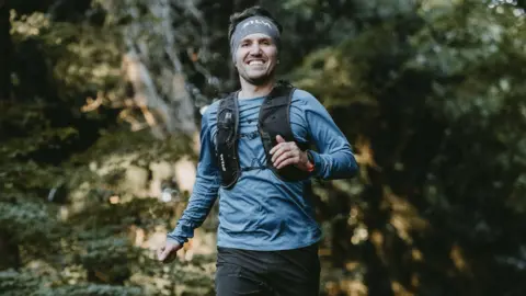 Sam Ellis Dan Beaumont. He is pictured smiling at the camera whilst running. He has short brown hair and a little facial hair. 