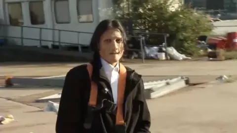Supplied A man with long black hair wearing a prosthetic on his face and dressed in a dark coat with a white shirt buttoned at the collar