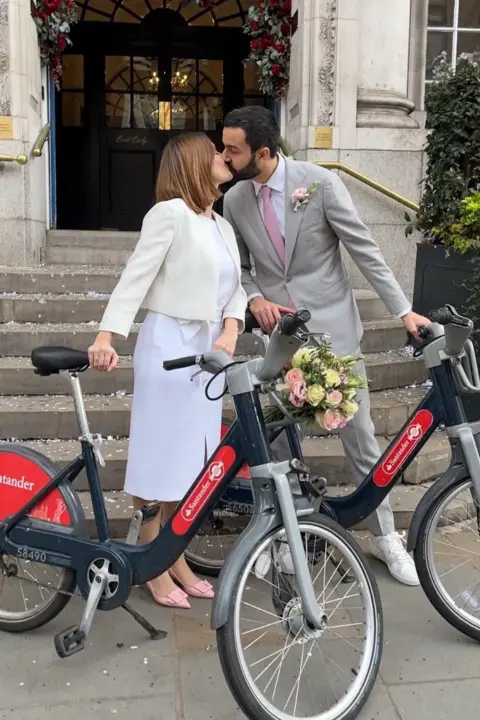 TfL A bride in a white dress and cropped jacket kisses a groom in a grey suit on the steps of Chelsea Old Town Hall. Both hold Santander Cycles beside them. Confetti is scattered on the steps, and an arch of red and white flowers decorates the doorway above.
