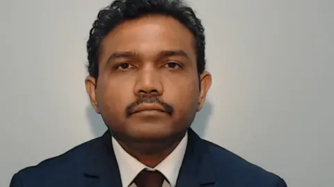 Vijay Gaikwad, wearing a dark blue suit and red tie with black hair and a black moustache, looks straight ahead in his police mughshot