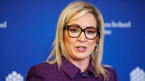 Michelle O'Neill. She has shoulder-length blonde hair, wearing glasses, and a purple jacket. A blue background is behind her.