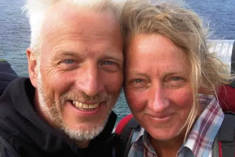 A woman, wearing a red jacket, poses for a selfie with her husband who has grey hair and is wearing a black jacket. In the distance is the sea. 
