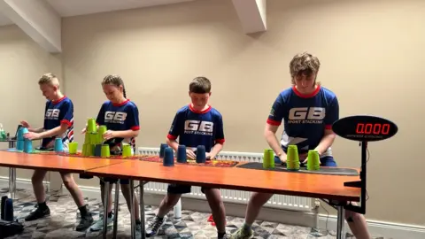 Four people stacking cups on a long table with a timer in front of them. They are all wearing blue and red jerseys.