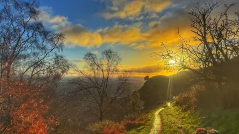 BBC Weather Watchers/Tarquin Shaw-Young The sun sets over a hill in the distance, turning the sky and clouds yellow. In the foreground are the bare branches of trees.