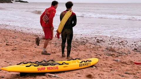 Two RNLI lifeguards with their backs to the camera stand by a yellow surfboard on a beach one wears red jacket and shorts while the other wears a black wetsuit