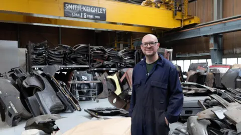 Bob Smith A man in blue overalls stands in a garage surrounded by car parts. Above his head is a yellow crane with a sign that reads Smith-Keighley Load 15 tons.