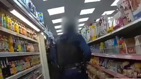 The back of a Met Police office raiding a shop. There are drinks in a fridge to the left and food items on shelving on the right.