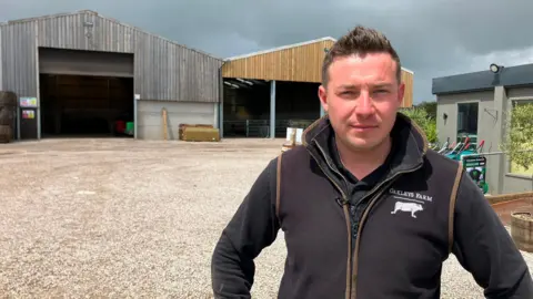 A man stands outside farm buildings, with barns and equipment in the background.
