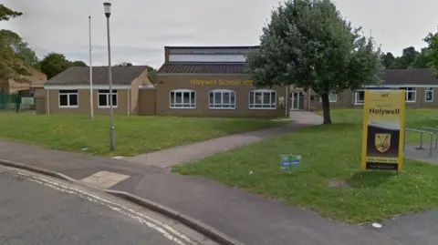 The front entrance of a school which has yellow signage saying "Holywell School". There is a pavement and a grass area in front of the building which is on one level. There is a street lamp on the left, a tree in the middle and bicycle racks on the right of the picture.