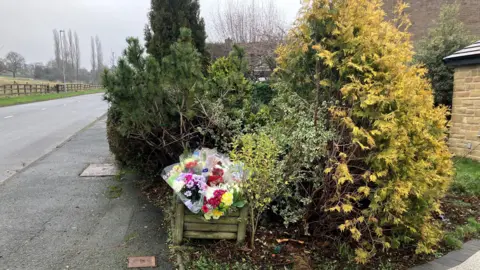 Steve Jones/BBC Flowers left at the scene of the crash. Also visible are some trees, some of which have been partially uprooted.