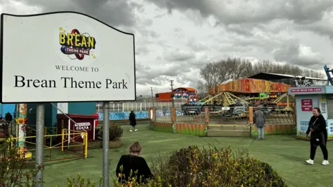 A large notice in the foreground saying 'Welcome to Brean Theme Park' and fairground rides in the background on a grass pitch with a few people milling around. The clouds are grey and gloomy