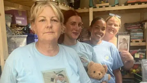 Dom Cotter Four women smile as they stand together in blue T-shirts. One is holding a teddy. They appear to be in a store room which is filled with bric-a-brac.