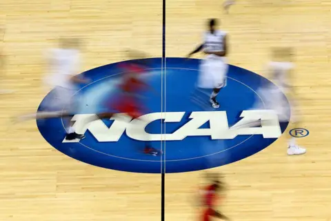 Blurry athletes pass over the NCAA logo on the floor of a basketball court