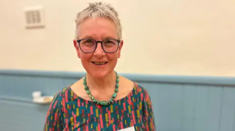 A woman with short, grey hair and glasses looks at the camera. She's wearing a multi-coloured top and a necklace of large green beads