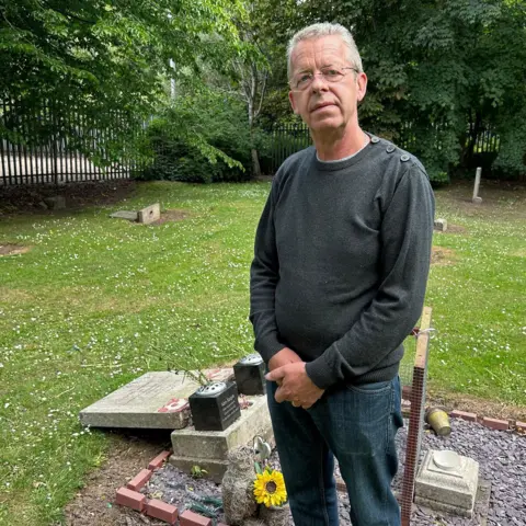 A man stands by a damaged grave site. He has a serious expression on his face. The grave is surrounded by short grass and other, standing headstones. The man wears a grey jumper, jeans and glasses.