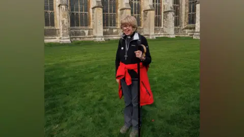 Neil Turner for Lambeth Palace Dame Sarah Mullally pictured outside Canterbury Cathedral ahead of her installation. Dame Sarah, who is stood outside the cathedral grounds, is wearing a black fleece, navy trousers, and has a red jacket tied around her waist. She is holding a black walking stick and is wearing a big cross around her neck. She is wearing walking boots. 