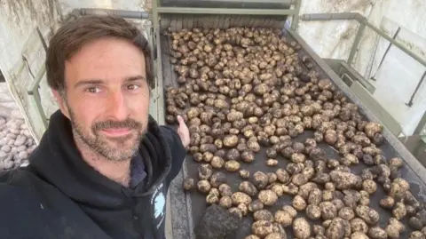 Luke Abblitt Luke Abblitt has a dark beard and moustache and is wearing a black hoodie, with the hood down. Behind him is a conveyor of potatoes straight from the field