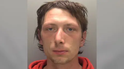 A police mugshot of a man with short dark brown hair wearing an orange sweatshirt. He is looking straight at the camera.